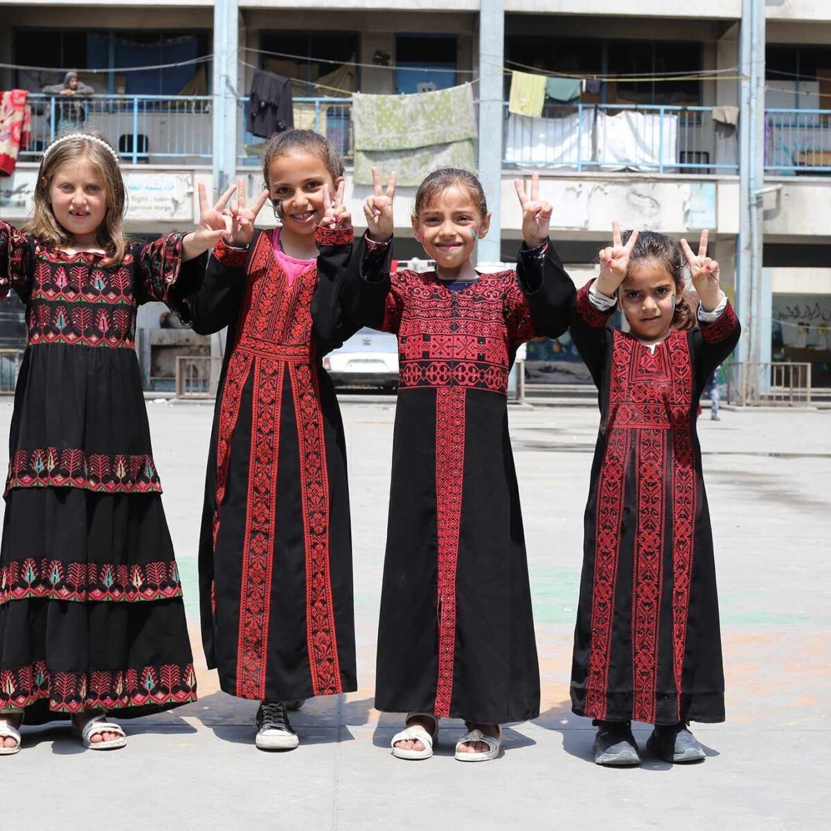 Four smiling girls in traditional Palestinian dress looking directly to camera making the peace sign with their hands